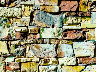 Colorful stone wall made up of various shapes and sizes of stones, arranged in an irregular yet harmonious pattern. The stones display a wide range of colors, including shades of yellow, brown, and green, with some stones showing patches of mixed hues. The texture of the stones is rough and uneven, adding a natural and rustic feel.