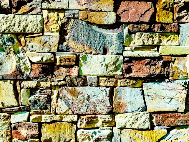 Colorful stone wall made up of various shapes and sizes of stones, arranged in an irregular yet harmonious pattern. The stones display a wide range of colors, including shades of yellow, brown, and green, with some stones showing patches of mixed hues. The texture of the stones is rough and uneven, adding a natural and rustic feel.