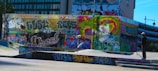 A vibrant urban skate park with walls covered in colorful graffiti art. A skateboarder is performing a trick on one of the ramps, wearing casual clothing and a cap. Another person is standing nearby, observing the scene. The environment is rich with dynamic street art, showcasing expressive lettering and abstract designs.