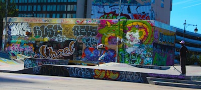 A vibrant urban skate park with walls covered in colorful graffiti art. A skateboarder is performing a trick on one of the ramps, wearing casual clothing and a cap. Another person is standing nearby, observing the scene. The environment is rich with dynamic street art, showcasing expressive lettering and abstract designs.