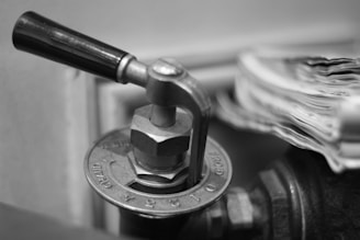 A close-up of a metal valve handle attached to a circular base with engraved numbers and markings. The handle is a lever style and is positioned horizontally. The image is in black and white, showing fine details of the metallic surfaces and textures.