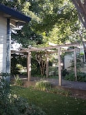 A wooden pergola structure stands in a garden, surrounded by lush green foliage and trees. The scene is bright and sunlit, with shadows cast by the leaves. The pathway beneath the pergola is bordered by various plants.