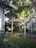 A wooden pergola structure stands in a garden, surrounded by lush green foliage and trees. The scene is bright and sunlit, with shadows cast by the leaves. The pathway beneath the pergola is bordered by various plants.
