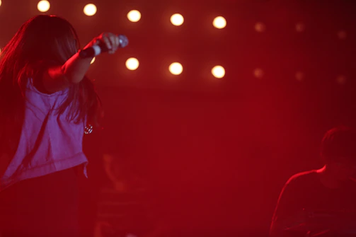 A close-up of a hand holding a microphone on stage with colorful stage lights blurred in the background.