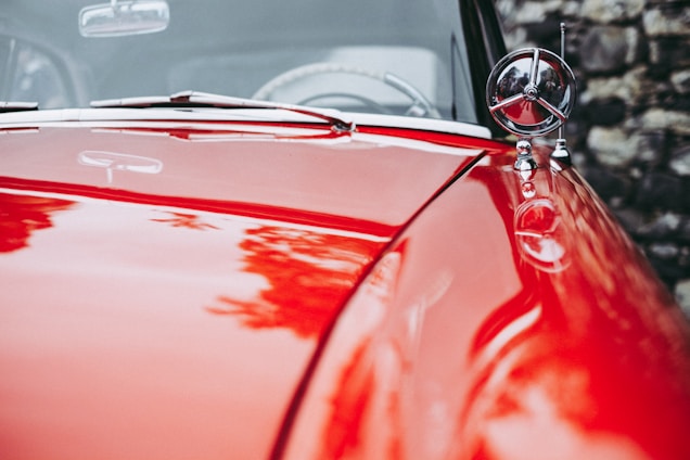 A polished red car hood with shiny chrome details and a circular ornament. The reflection of surrounding foliage is visible on the surface, and a blurred stone wall can be seen in the background.
