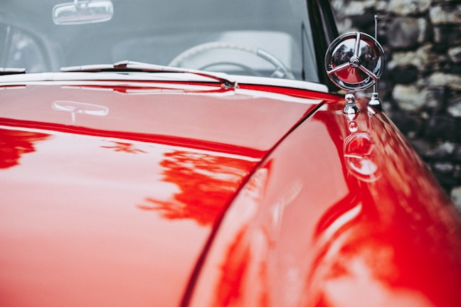 A polished red car hood with shiny chrome details and a circular ornament. The reflection of surrounding foliage is visible on the surface, and a blurred stone wall can be seen in the background.