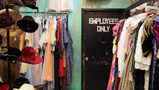 A room filled with a variety of clothing items hanging on racks, with colorful dresses and garments on both sides. To the left, there is a display of hats, featuring several different colors and styles. In the center back of the room is a closed black door labeled 'EMPLOYEES ONLY'. The wall behind the clothing has a teal color.