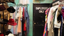 A room filled with a variety of clothing items hanging on racks, with colorful dresses and garments on both sides. To the left, there is a display of hats, featuring several different colors and styles. In the center back of the room is a closed black door labeled 'EMPLOYEES ONLY'. The wall behind the clothing has a teal color.
