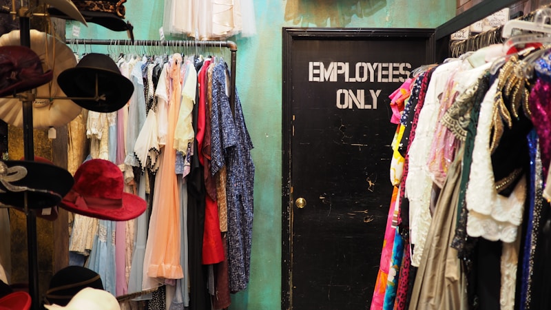 A room filled with a variety of clothing items hanging on racks, with colorful dresses and garments on both sides. To the left, there is a display of hats, featuring several different colors and styles. In the center back of the room is a closed black door labeled 'EMPLOYEES ONLY'. The wall behind the clothing has a teal color.