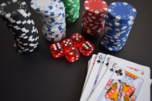 Stacks of poker chips in various colors including black, white, red, blue, and green are accompanied by red dice and a hand of playing cards featuring face cards such as the Queen and King of Clubs.