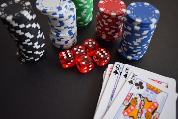 Stacks of poker chips in various colors including black, white, red, blue, and green are accompanied by red dice and a hand of playing cards featuring face cards such as the Queen and King of Clubs.