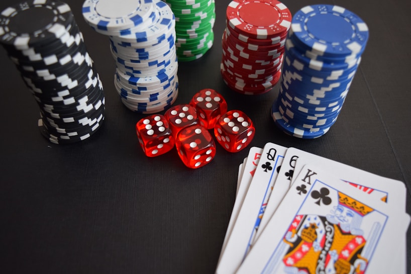 Stacks of poker chips in various colors including black, white, red, blue, and green are accompanied by red dice and a hand of playing cards featuring face cards such as the Queen and King of Clubs.