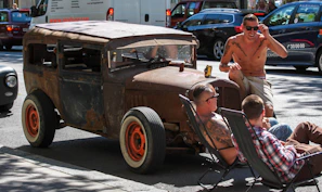 A group of friends in various V8 Clothing outfits gathered around a roaring V8 engine, laughing and enjoying a sunny day.