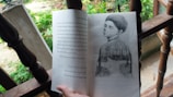 An open book resting on a wooden surface features an illustration of a contemplative figure with closed eyes, blended with a nature scene. The background suggests an outdoor setting with greenery and leaves.