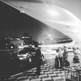 A black and white photograph captures a nighttime scene outside of an airport or transport terminal. Several people stand on a patterned sidewalk, some with luggage, under a large, curved architectural canopy. Cars and taxis are on the road nearby, with city buildings visible in the background. The setting is illuminated by outdoor lighting.