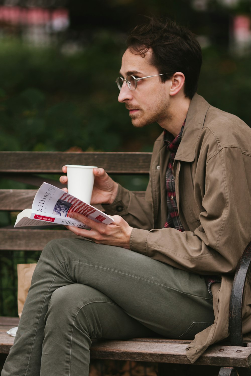 갈색 가죽 재킷과 회색 데님 청바지를 입은 남자가 벤치에 앉아 책을 읽고 있습니다.