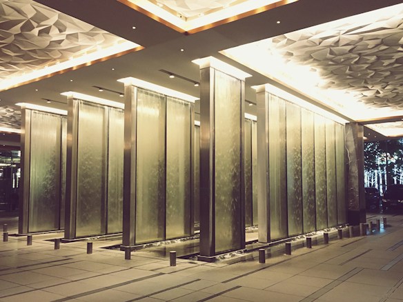 Several tall, rectangular water fountains arranged in a symmetrical manner in an indoor space with a modern, geometric ceiling and subtle lighting. The floors and walls are sleek and clean, featuring a blend of metal and stone textures.