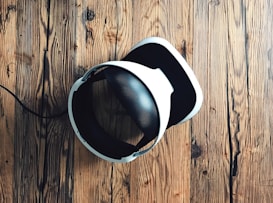 A virtual reality headset with a sleek design rests on a rustic wooden surface, creating a contrast between modern technology and natural textures. The headset is mainly black and white.