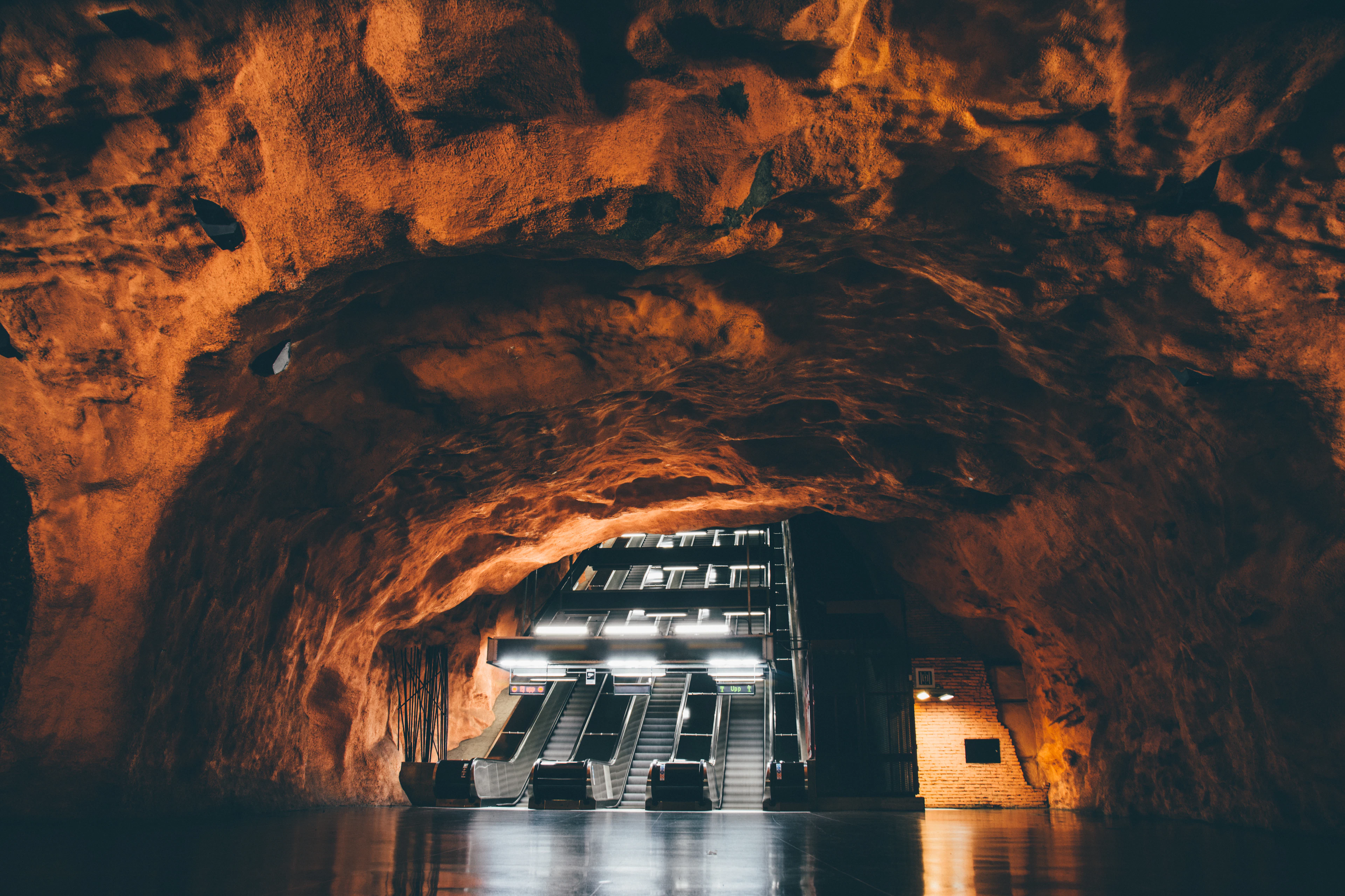 Escalators rising into a vast blasted-bedrock cavern in the Stockholm metro, the rough rock ceiling glowing orange above a polished platform floor.