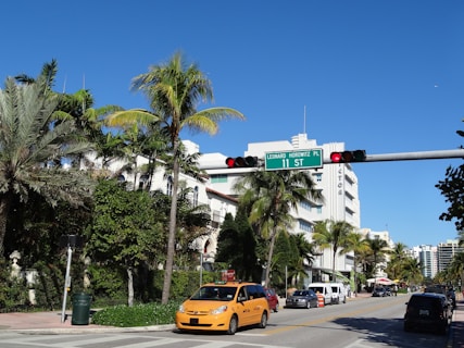 A street scene featuring palm trees and art deco buildings under a clear blue sky. A yellow taxi is prominently visible, waiting at a traffic light. The intersection is marked by a street sign with palm trees and various vehicles lining the road.
