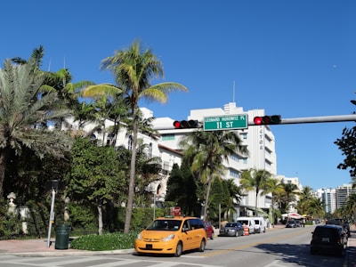 A street scene featuring palm trees and art deco buildings under a clear blue sky. A yellow taxi is prominently visible, waiting at a traffic light. The intersection is marked by a street sign with palm trees and various vehicles lining the road.