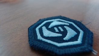 A round embroidered patch with a white and blue color scheme lies on a wooden surface. The patch features a design of two hands reaching towards each other, enclosed within a blue border.