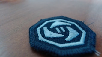 A round embroidered patch with a white and blue color scheme lies on a wooden surface. The patch features a design of two hands reaching towards each other, enclosed within a blue border.