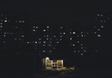 A small, illuminated kiosk is situated in front of a dimly lit urban apartment complex at night. The kiosk is well-lit, displaying various goods with a warm glow, while the surrounding buildings are mostly dark with scattered lights in the windows.