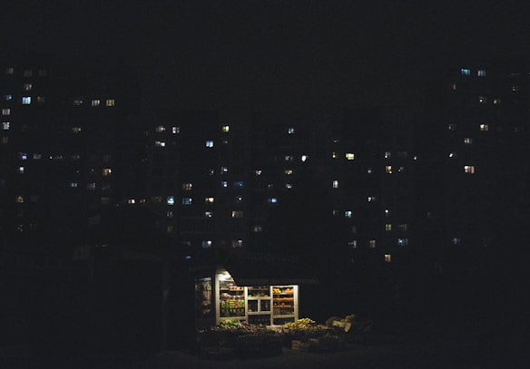 A small, illuminated kiosk is situated in front of a dimly lit urban apartment complex at night. The kiosk is well-lit, displaying various goods with a warm glow, while the surrounding buildings are mostly dark with scattered lights in the windows.
