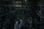 A dimly lit warehouse containing shelves stacked with various mechanical parts, possibly car engines or machinery components. The parts appear to be arranged in a cluttered manner with pipes and wires visible. Two covered figures stand in the foreground, which might be statues or objects draped with cloth.
