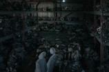 A dimly lit warehouse containing shelves stacked with various mechanical parts, possibly car engines or machinery components. The parts appear to be arranged in a cluttered manner with pipes and wires visible. Two covered figures stand in the foreground, which might be statues or objects draped with cloth.