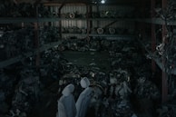 A dimly lit warehouse containing shelves stacked with various mechanical parts, possibly car engines or machinery components. The parts appear to be arranged in a cluttered manner with pipes and wires visible. Two covered figures stand in the foreground, which might be statues or objects draped with cloth.