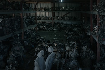 A dimly lit warehouse containing shelves stacked with various mechanical parts, possibly car engines or machinery components. The parts appear to be arranged in a cluttered manner with pipes and wires visible. Two covered figures stand in the foreground, which might be statues or objects draped with cloth.