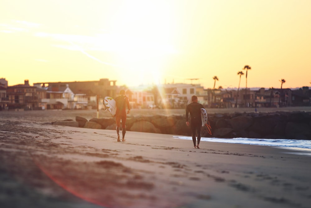 two person walking on seashore while holding surfboards