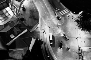 An aerial view of a city intersection at night with cars and a bus navigating the streets. The streets are well-lit, and there is a clear view of crosswalks and the shadows they cast. Buildings line the streets with distinct geometric shapes and patterns on the rooftops.