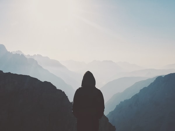 A hooded figure staring at clusters of mountains shrouded in fog.