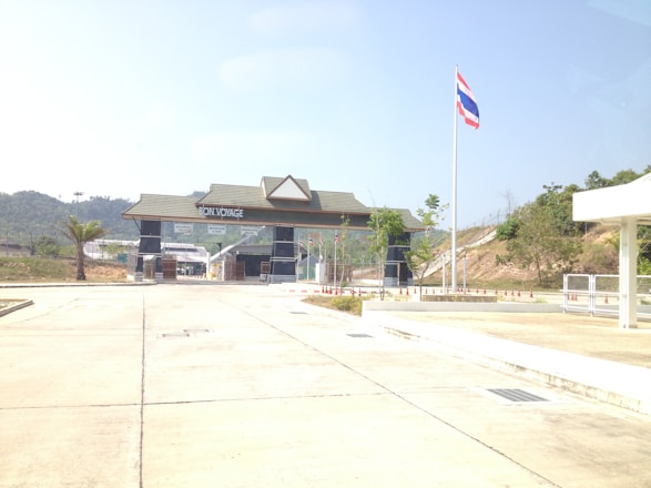 A border checkpoint with a large gate structure displaying the words 'Bon Voyage'. The gate is situated on a paved area, with a clear blue sky above. A flagpole with the Thai flag is prominently visible, and there are small traffic cones arranged in front of the building. The landscape surrounding the checkpoint includes gentle hills and sparse greenery.
