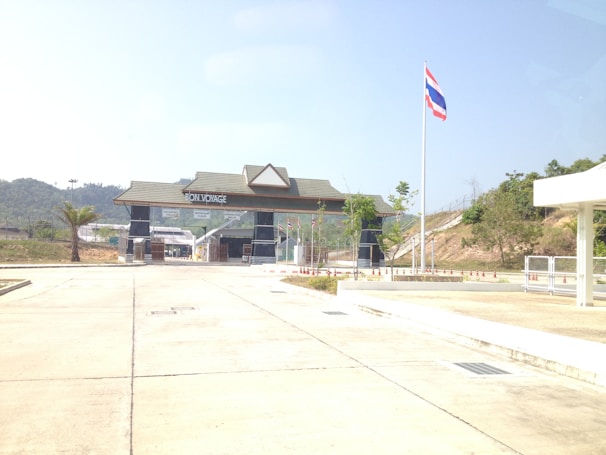 A border checkpoint with a large gate structure displaying the words 'Bon Voyage'. The gate is situated on a paved area, with a clear blue sky above. A flagpole with the Thai flag is prominently visible, and there are small traffic cones arranged in front of the building. The landscape surrounding the checkpoint includes gentle hills and sparse greenery.