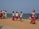 A group of children in school uniforms walk along a sandy path near the ocean, accompanied by two adults who may be their guardians or teachers. The children carry red school bags and appear to be walking with purpose, possibly towards or from school. The setting is bright and open, with the sea visible in the background, and the sky is clear.