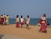 A group of children in school uniforms walk along a sandy path near the ocean, accompanied by two adults who may be their guardians or teachers. The children carry red school bags and appear to be walking with purpose, possibly towards or from school. The setting is bright and open, with the sea visible in the background, and the sky is clear.