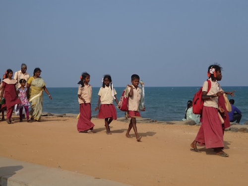 A group of children in school uniforms walk along a sandy path near the ocean, accompanied by two adults who may be their guardians or teachers. The children carry red school bags and appear to be walking with purpose, possibly towards or from school. The setting is bright and open, with the sea visible in the background, and the sky is clear.