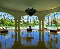 A spacious, open-air pavilion with elegant columns and a large chandelier hanging from the ceiling. The floor is shiny and reflects the architecture and furniture. Outside, lush greenery and palm trees surround a tranquil body of water.