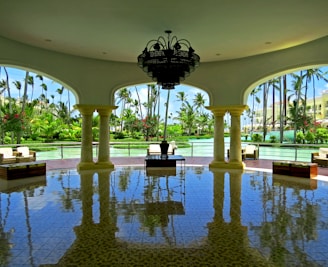 A spacious, open-air pavilion with elegant columns and a large chandelier hanging from the ceiling. The floor is shiny and reflects the architecture and furniture. Outside, lush greenery and palm trees surround a tranquil body of water.