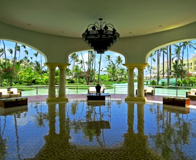 A spacious, open-air pavilion with elegant columns and a large chandelier hanging from the ceiling. The floor is shiny and reflects the architecture and furniture. Outside, lush greenery and palm trees surround a tranquil body of water.