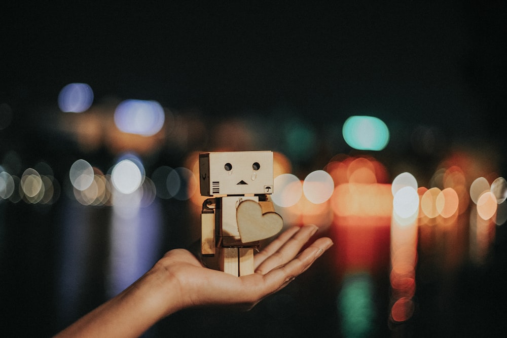 A person's hand holding up a box character with a heart, in front of a blurred out city background.