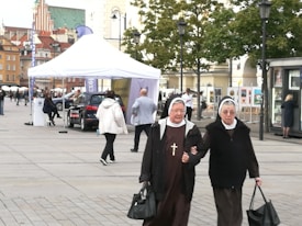 Two nuns dressed in traditional habits, holding hands and walking on a paved square. They are carrying black bags and seem to be engaged in conversation. In the background, there's a busy street scene with a white tent, cars, people walking, and historic buildings with a green-domed structure visible.