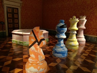 Several oversized chess pieces, each painted in a unique and colorful pattern, are arranged on a wooden patterned floor in a richly decorated room. The walls are adorned with red and gold ornate wallpaper. A large door with decorative panels is visible in the background, and a pink and green object resembling a fountain or a piece of modern art is positioned to the side.