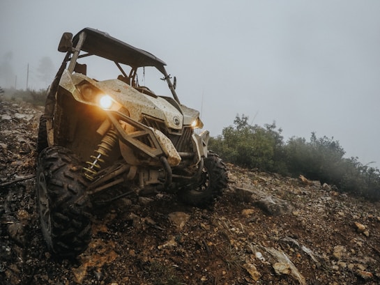 A rugged ATV with prominent off-road tires and headlights turned on navigates a rocky and muddy trail on a foggy day. The vehicle is situated at an angle, highlighting its suspension system, with dense greenery visible in the background.