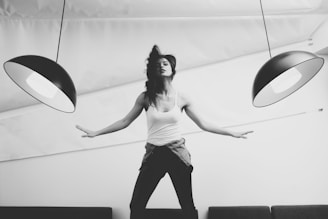 A person poses dynamically with arms outstretched beneath two large, modern pendant lamps, creating a sense of balance and symmetry. The image is in black and white, adding a timeless and artistic feel. The person is wearing casual clothing, including a tank top and a shirt tied around the waist.