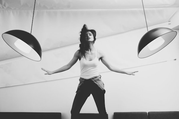 A person poses dynamically with arms outstretched beneath two large, modern pendant lamps, creating a sense of balance and symmetry. The image is in black and white, adding a timeless and artistic feel. The person is wearing casual clothing, including a tank top and a shirt tied around the waist.